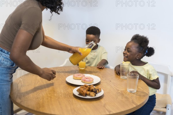 Mother pouring orange juice for her two children during breakfast, with donuts and pastries on the