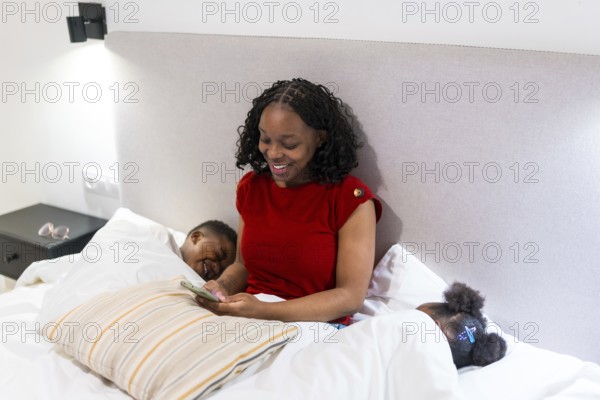 Smiling mother using smartphone while her two children are sleeping in bed, enjoying a quiet moment