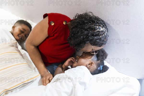 Mother kisses her daughter goodnight while putting her to bed, as her brother watches from the