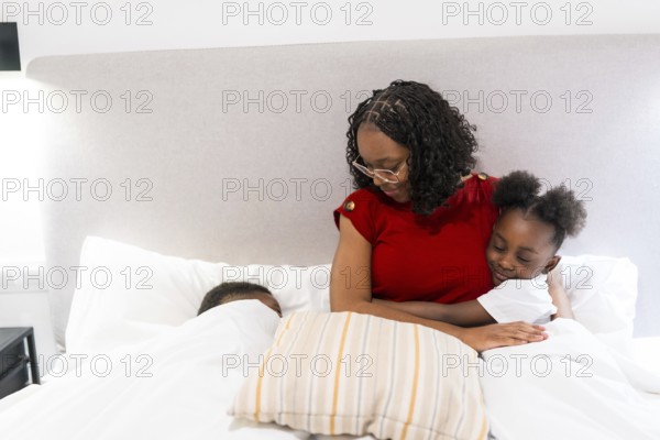 Young mother embracing her daughter in bed, while her son peacefully sleeping beside them, creating