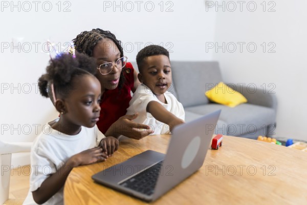 Young mother guiding her two children while using a laptop on a wooden table at home, fostering