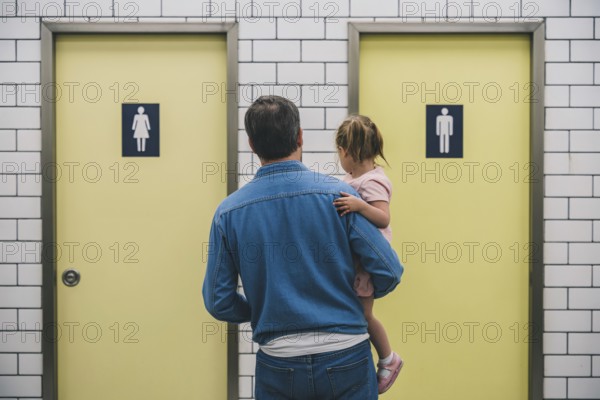 Father with young daughter hesitating in front of men's and women's restroom doors. Concept of