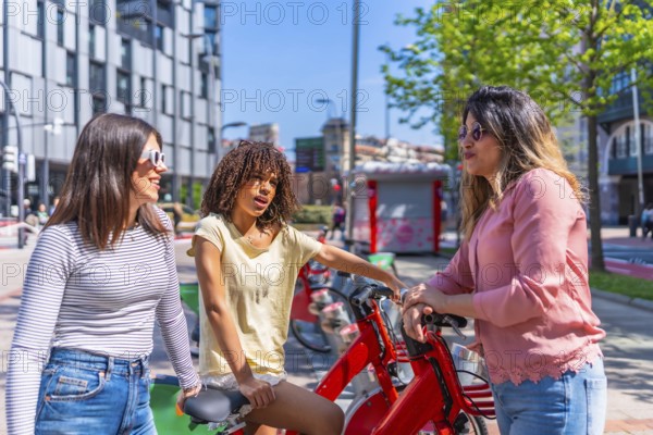 Three young women chatting happily near a bike sharing station in a vibrant city, enjoying a sunny