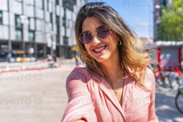 Smiling young woman wearing trendy sunglasses, capturing a joyful selfie in a vibrant city center