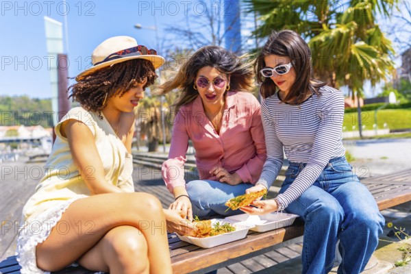 Three female friends enjoying a pizza lunch together on a park bench during a sunny summer day