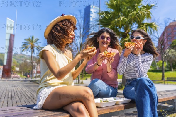 Three young women savoring delicious pizza while sitting on a bench in a vibrant city park,