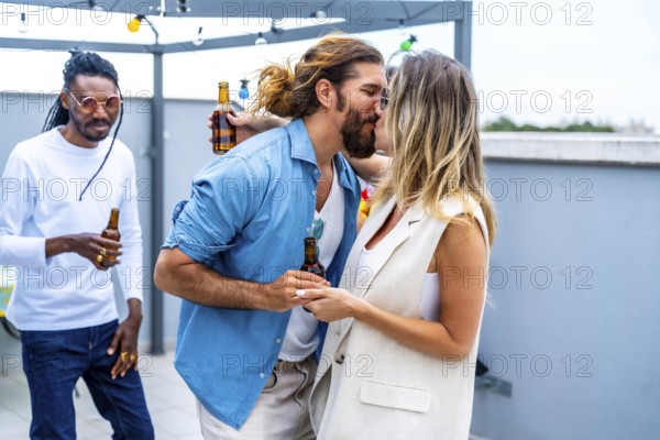 Young couple kissing and holding hands at rooftop party while friends drinking beer and having fun