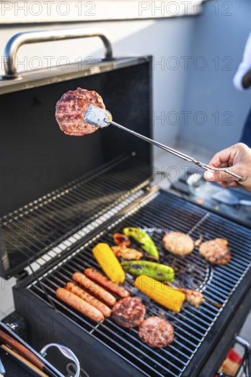 Hand using tongs to grill hamburgers, sausages, corn and peppers on a barbecue grill during a