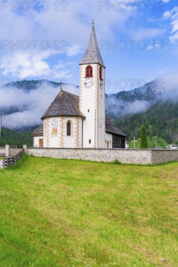 Church with clock tower in a green alpine landscape under a cloudy sky, Dolomites, South Tyrol,