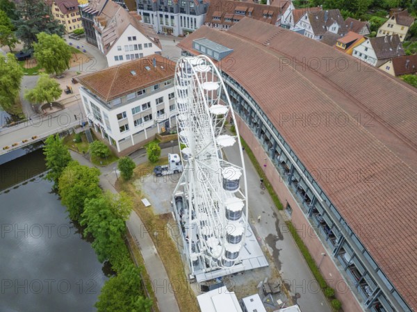 A large white Ferris wheel stands next to modern buildings with red roofs in an urban setting,