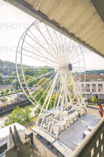 A Ferris wheel is set up in a town next to a river, surrounded by green landscape and buildings,