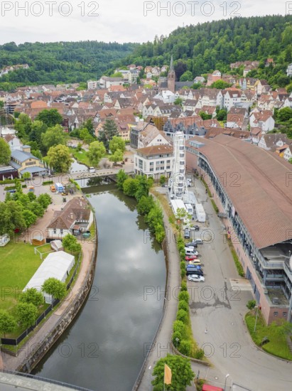 A river flows through a town with a Ferris wheel, surrounded by historic buildings and green hills,