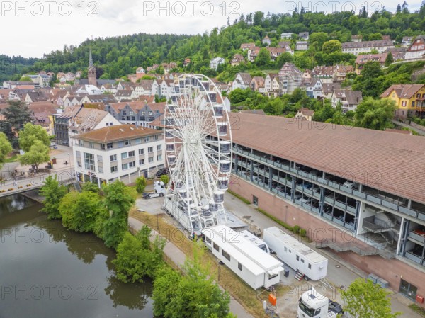 A Ferris wheel next to urban buildings embedded in a summer landscape with wooded hills, Ferris