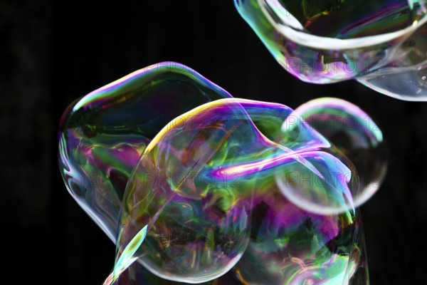 Large, irregular, iridescent soap bubbles in the colours of the rainbow against a black background,