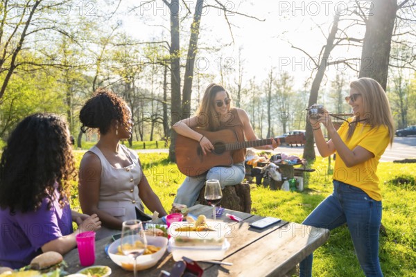 Four female friends enjoying a picnic in a park, with one playing guitar, another taking photos,
