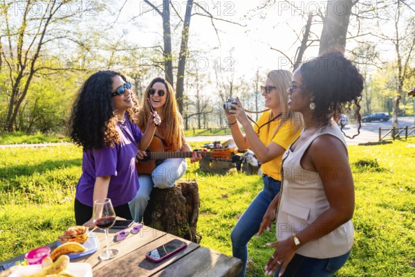 Four cheerful female friends are having a picnic in a sunny park, playing guitar, taking photos,
