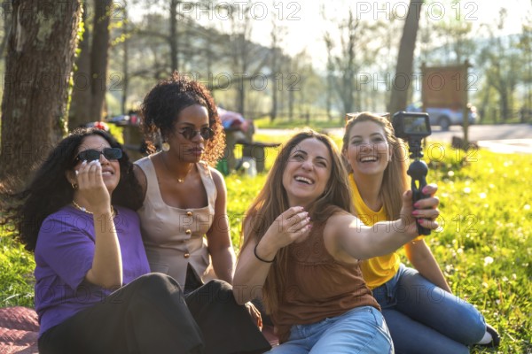 Four multi ethnic female friends are sitting on grass in a park, taking a selfie with an action