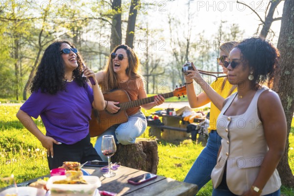 Four female friends enjoying a picnic in a park, singing, playing guitar, taking photos, and having