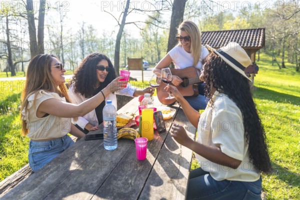 Happy multi ethnic women enjoying a picnic in nature, toasting with drinks, playing guitar, and