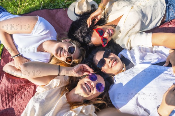Four diverse young women lying together in a park on a blanket wearing sunglasses and smiling up at