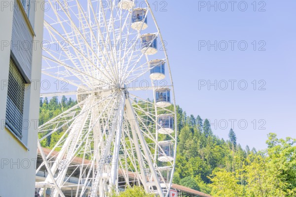 A large Ferris wheel stands next to a building with a wooded hill and clear sky in the background,
