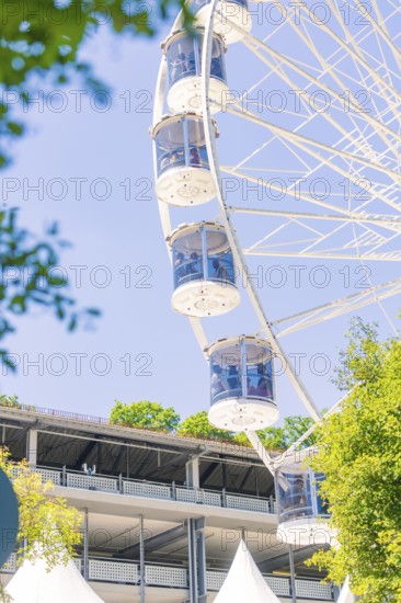The Ferris wheel with glass cabins rises next to a large building surrounded by sunny trees, 950