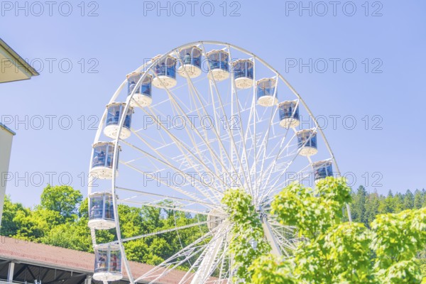 A white Ferris wheel turns in front of a building surrounded by green vegetation on a clear summer