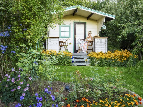 A woman in a dress sits relaxed on the small veranda of a garden shed and reads. Blooming summer