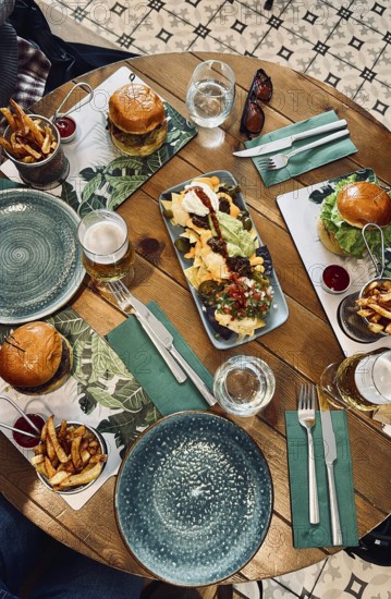 Top view of a wooden restaurant table with nachos, burgers, french fries, and beer, suggesting a