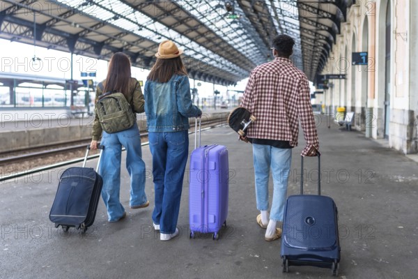 Three young tourists are walking away from camera in a train station with their suitcases and a