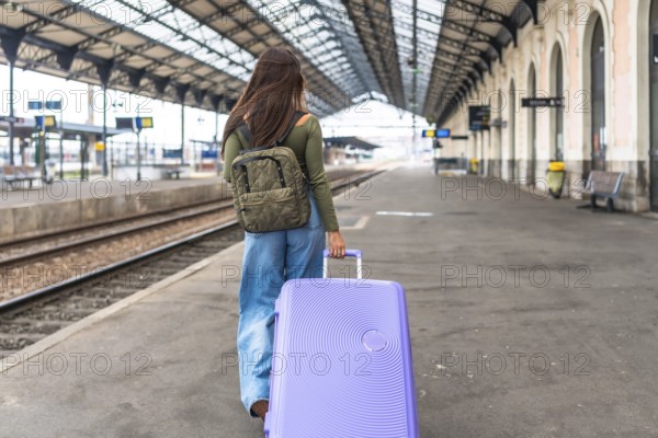 Tourist walking on railway platform pulling trolley suitcase and wearing backpack, traveling by