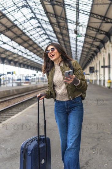 Happy female tourist walking through a bustling train station, using her smartphone while pulling a