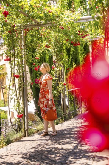 Woman in a red dress walks through a flowering garden pavilion, surrounded by green foliage,