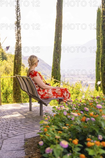 Woman in a red dress sitting on a bench surrounded by flowers and cypresses, Botanical Garden, The