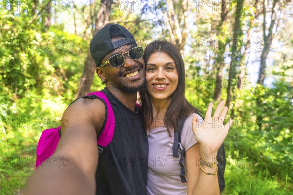 Interracial couple smiling and waving while taking a selfie during a vibrant hike in a lush, green