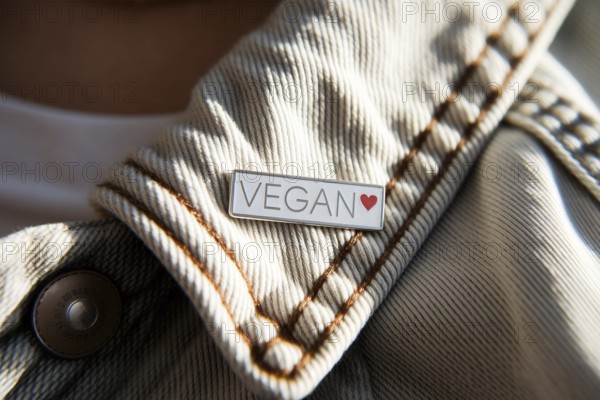 Close-up of silver pin with VEGAN text and red heart on worn cotton jacket. Symbol of ethical