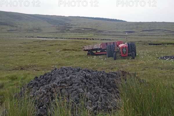 Old red tractor standing in boggy fields against a hilly backdrop with grey sky, peat cutting, Isle