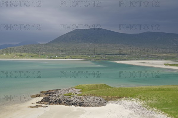Wide sandy beach meets turquoise waters, surrounded by green hills and mountains under a gloomy