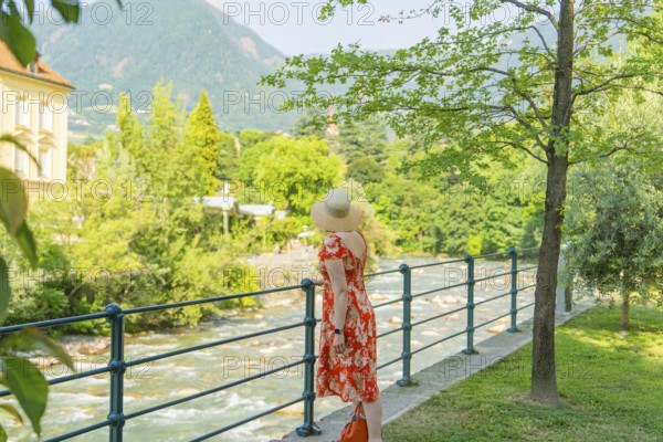 A woman looks at a river in the middle of a green, summery landscape, Merano, Italy