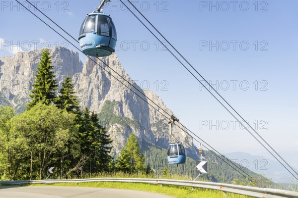 Two blue cable car gondolas hovering over trees in front of a rocky mountain range, Alpe di Siusi,