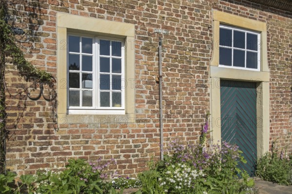 A rustic brick wall with two windows, wooden door and flowering plants, Haus Welbergen, Ochtrup,
