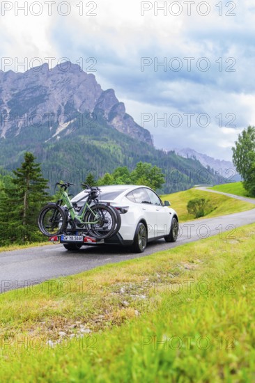 White car with bicycles on a road in a mountainous, green landscape with cloudy sky, electric car