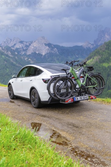 White car with bicycles on wet road, surrounded by mountains and rain clouds, electric car on