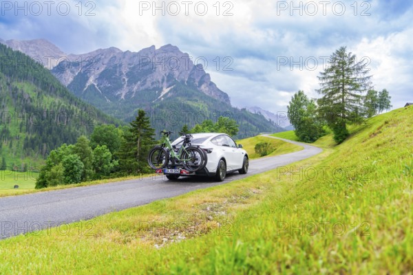 White car with bicycles on a road through green, hilly landscape with cloudy sky, electric car on
