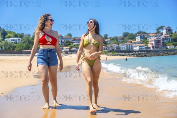 Two cheerful young women wearing bikinis and sunglasses are enjoying a walk on a sunny beach,