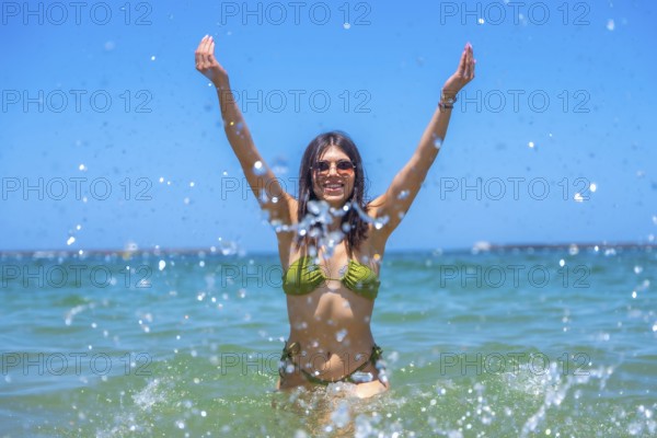 Young woman wearing sunglasses and a green bikini is raising her arms and splashing water in the