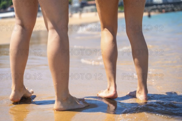 Two women standing barefoot on the wet sand of a sunny beach, enjoying the warm water and the
