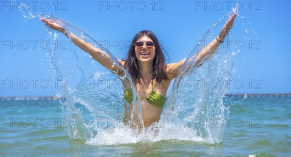Young woman wearing stylish sunglasses and a vibrant bikini joyfully splashing in the clear ocean