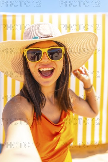 Young woman wearing sunglasses and a straw hat taking a selfie while smiling at the camera on a