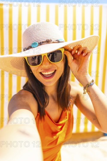 Young woman taking a selfie on a sunny beach, wearing an orange dress and a wide brimmed sun hat